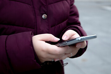 Young girl with a mobile device in her hands typing, wears maroon winter clothes while on the street.