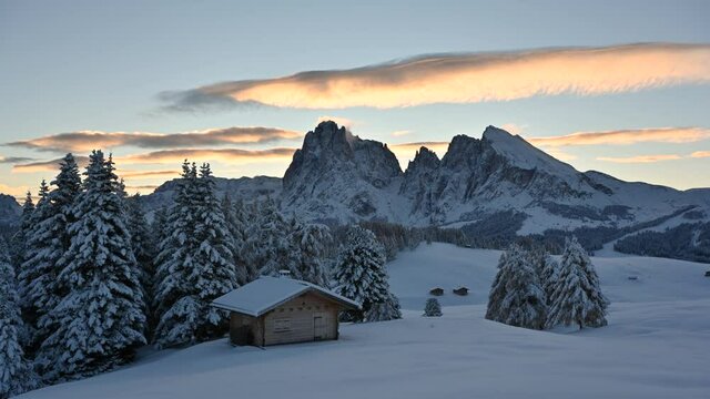 Timelapse footage with small wooden log cabin on meadow Alpe di Siusi on winter time. Seiser Alm, Dolomites, Italy. Snowy hills with orange larch and Sassolungo and Langkofel mountains group. 4k video