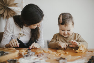 Adorable happy daughter and mother making together christmas cookies on messy table. Cute toddler girl helper with mom cutting dough for gingerbread cookies. Family preparations for xmas holidays