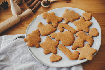 Delicious baked gingerbread cookies on modern plate on wooden table with napkin, rolling pin, spices. Making Christmas cookies close up. Holiday preparation, xmas culinary. Biscuits recipe