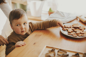 Cute little girl grabbing freshly baked gingerbread cookie on wooden table in modern room. Adorable...