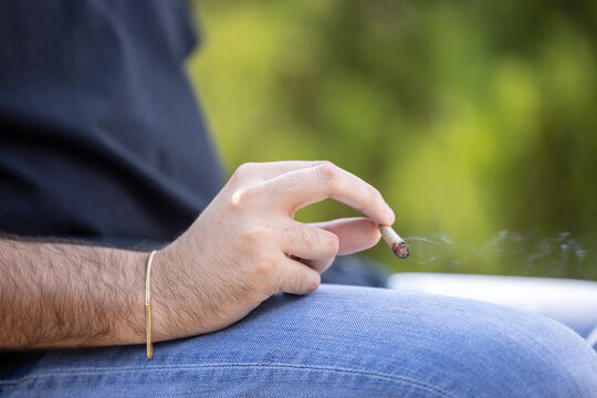 Detail of man holding a lit CBD pre-roll joint, cigarette with smoke rising sitting outdoors