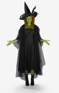 Full length portrait on white of a woman with green body paint dressed up in a witch costume for Halloween with her arms out.
