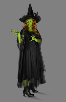 Portrait of a woman dressed as a witch against a gray background with green body paint, looking off camera with her hands up casting a spell.