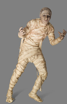 Man dressed up as a mummy for Halloween, making a stiff movement away from camera, against a gray background