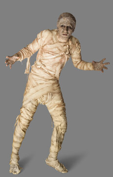 Man dressed up as a mummy for Halloween making stiff arm movements against a gray background