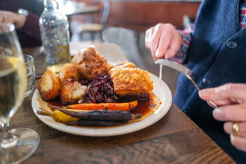 The golden thick crackling on top of roast pork belly on a white plate with roast potatoes and Yorkshire pudding with vegetables in a restaurant. A mans hands can be seen holding a knife and fork.