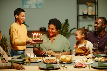 Portrait of smiling teenage boy bringing Birthday cake to happy African-American grandmother celebrating with family