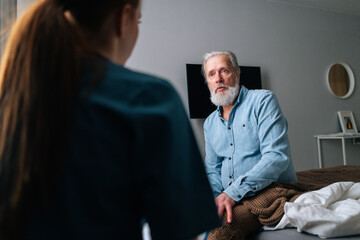 Close-up view from back of female doctor talking to sick senior male patient sitting on bed at...