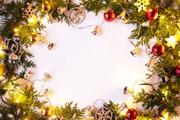 Fototapeta na wymiar Christmas or New Year background: fir branches on a white background with wooden toys and a sparkling garland, bells, balls. White frame for congratulations