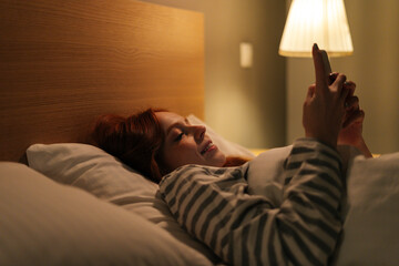 Close-up of cheerful young woman using smartphone, looking on screen, typing online message on social media, lying on bed late at night. Tired insomnia lady browsing smartphone at night in bed.