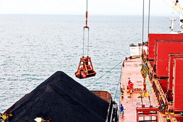 Loading coal from cargo barges onto a bulk carrier using ship cranes and grabs at the port of Muara...