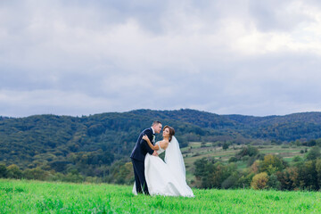Newlyweds are walking in the field on the green grass. A man hugs, tilts a woman, against the backdrop of beautiful nature.