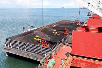 Loading coal from cargo barges onto a bulk carrier using ship cranes and grabs at the port of Muara...