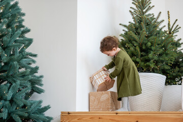 Little girl in green dress stands on the wooden steps by the Christmas tree, opens boxes with gifts