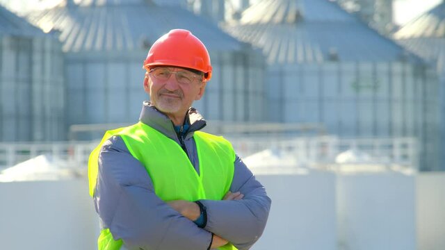 Grain elevator port storage. Bearded worker man in orange hardhat stands with arms crossed at construction site looking at built modern complex