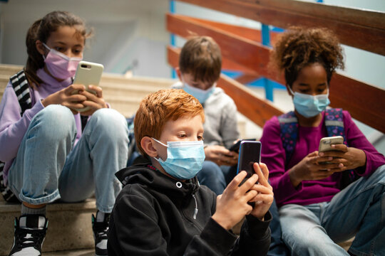 Group of school children wearing face masks and using smart mobile phones before class.