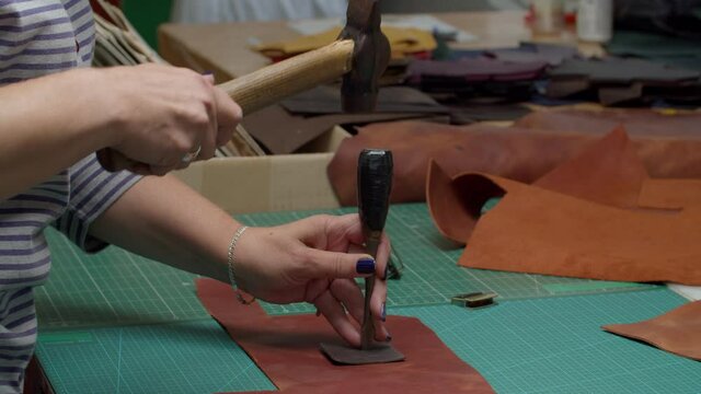 Midsection of unrecognizable female worker making notches with hummer and metal puncher indoors. Process of making genuine leather detail in handmade workshop