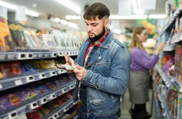 Focused bearded guy choosing chocolate on shelves in grocery store