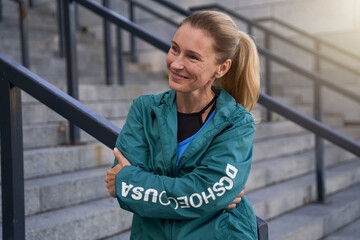 Portrait of cheerful middle aged sportswoman in sportswear smiling aside while standing outdoors after training in the city