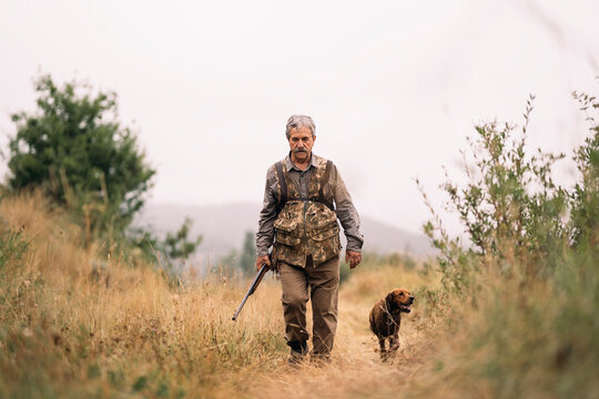 Mature hunter man holding a shotgun and walking through a field 