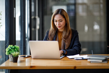 Fototapeta premium Asian young businesswoman sitting at a table with laptop computer and doing math finance report on wooden desk, tax, accounting, statistics and analytical research concept
