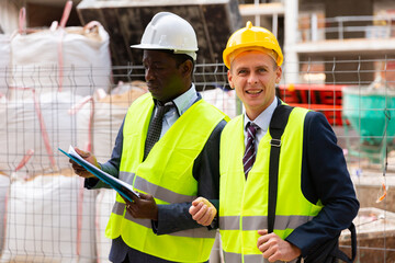 Two architects, Caucasian and African-american men in warnvests, standing on building site. One of them smiling and looking in camera.