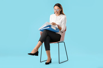 Beautiful young woman with folders sitting on chair against color background