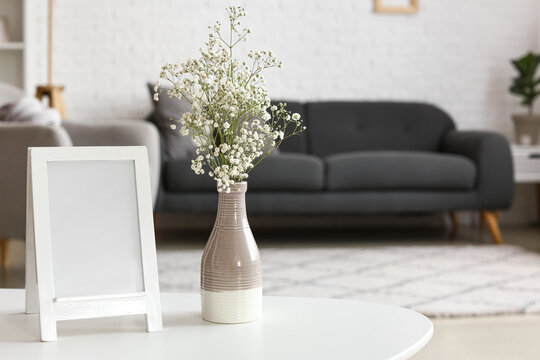 Vase with gypsophila flowers and blank frame on table in living room