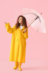 Young African-American woman wearing yellow raincoat and gumboots with umbrella on pink background