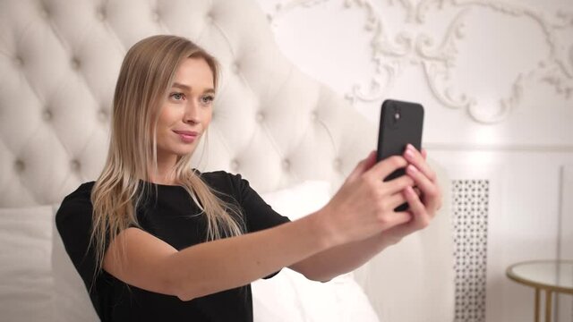 Charming young adult woman poses on bed in black shirt to take cute picture, photos with phone for social media likes. Casual lady touches smartphone screen to record, film video of herself in bedroom