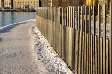 Snowy walking path at Stockholm bay on sunny freezing winter day with steaming water, wooden Scandinavian fence in front and city buildings in a background. Winter time in capital of Sweden  