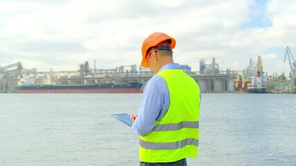 Sea industry port ship safety. Concentrated manager with orange helmet inputs checkup results on tablet inspecting equipment in dockyard