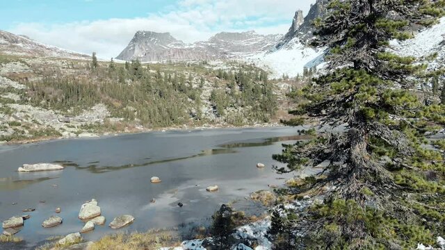 Sleeping Sayan rocky and taiga behind the Raduzhnoe lake. Snow lies, the sky is blue, stones and cold water. Ergaki park