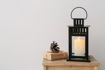 Black lantern with burning candle, gift and fir cone on stool near light wall