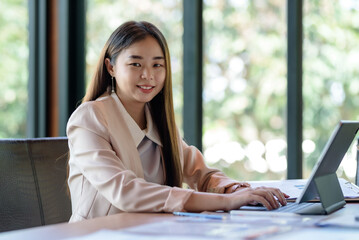 Successful smiling beautiful young Asian businesswoman sitting with laptop and computer while doing some paperwork at the office. Looking at camera.