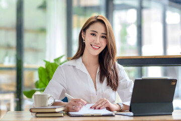 Successful smiling beautiful young Asian businesswoman sitting with laptop and computer while doing some paperwork at the office. Looking at camera.