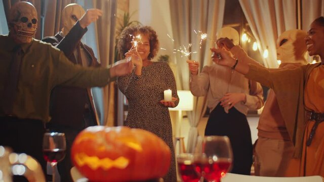 Rack Focus Shot Of Cheerful Masked Friends Dancing With Sparklers At Halloween Home Party There Is Jack-o-lantern On Dinner Table