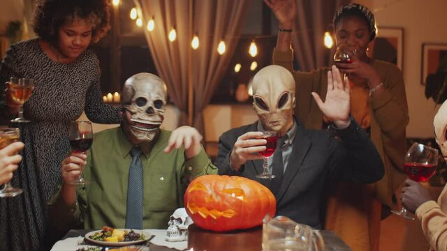 Company of friends in Halloween masks clinking wine glasses in toast, drinking and dancing at home dinner party