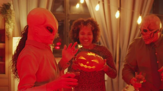 Group of young friends in scull and alien masks holding wine glasses and jack-o-lantern while dancing together at home Halloween party