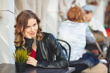 Young beautiful woman is sitting in a summer cafe. Curly blonde in a black leather jacket sits at a table. Looks into camera