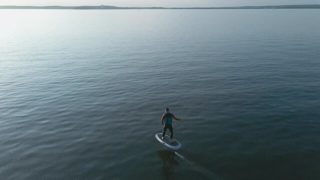 Man riding on a hydrofoil surfboard on large blue lake in sunny weather. Aerial shot of man riding a hydrofoil surfboard in the middle of lake. High quality 4k footage