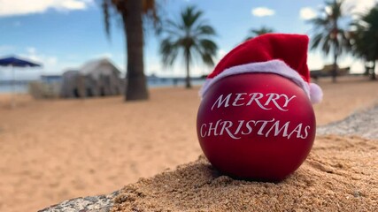 Christmas, Red bomb in Santa's hat on the exotic beach with palm trees and blue sky on the background with clouds. Hawaii, Canary islands, Bali, Thailand. Merry christmas from paradise. 