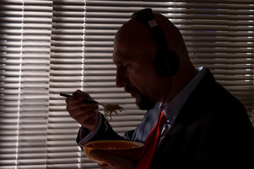 A man in a suit and headphones eats pasta against the background of a window with blinds. A special agent's lunch at work, wiretapping conversations.