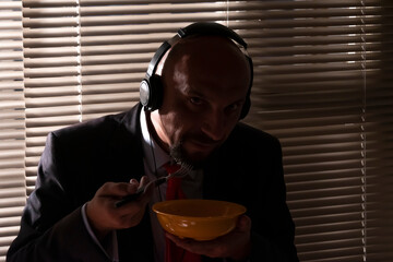 A man in a suit and headphones eats pasta against the background of a window with blinds. A special agent's lunch at work, wiretapping conversations. - obrazy, fototapety, plakaty