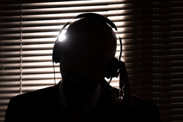 An FBI secret agent listens with headphones and records a conversation against the background of a window with blinds, silhouette lighting, selective focus, dark tone.