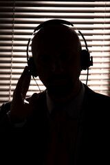 An FBI secret agent listens with headphones and records a conversation against the background of a window with blinds, silhouette lighting, selective focus, dark tonality. - obrazy, fototapety, plakaty