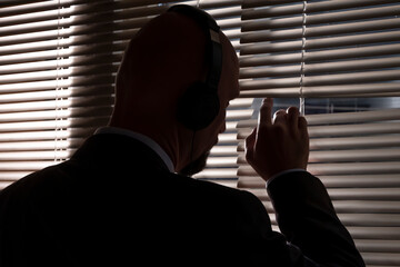 A secret agent eavesdrops on a conversation with headphones, looks through the window blinds at the street, , silhouette illumination, selective focus, dark tone. - obrazy, fototapety, plakaty