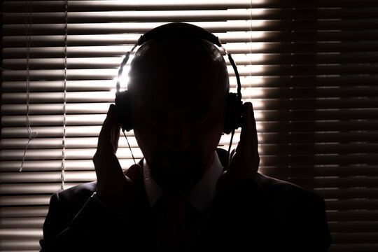 An FBI secret agent listens with headphones and records a conversation against the background of a window with blinds, silhouette lighting, selective focus, dark tone.