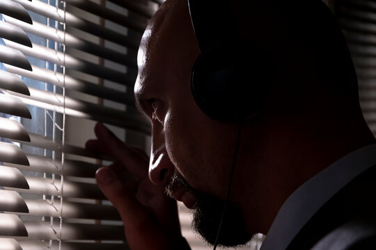 A secret agent eavesdrops on a conversation with headphones, looks through the window blinds at the street, , silhouette illumination, selective focus, dark tone.